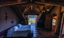 A cozy wooden cabin interior with a bed facing an open door leading to a balcony. The balcony offers a serene view of a lake surrounded by trees in the background. The room features wooden beams, a ceiling fan, and a warm, rustic decor.