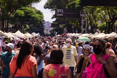 A crowded street market with numerous people walking between stalls on either side. The market is set under lush green trees, and many individuals are wearing summer clothing. Some people are wearing face masks, suggesting a concern for health safety. The atmosphere appears lively and bustling, with a variety of stalls featuring striped canopies visible in the background.