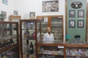 An old pharmacy with wooden display cabinets filled with various bottles and containers. A mannequin wearing a white lab coat stands behind the counter. Several framed photographs and vintage advertisements adorn the walls. The atmosphere invokes a sense of nostalgia and history.