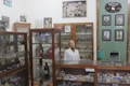 An old pharmacy with wooden display cabinets filled with various bottles and containers. A mannequin wearing a white lab coat stands behind the counter. Several framed photographs and vintage advertisements adorn the walls. The atmosphere invokes a sense of nostalgia and history.