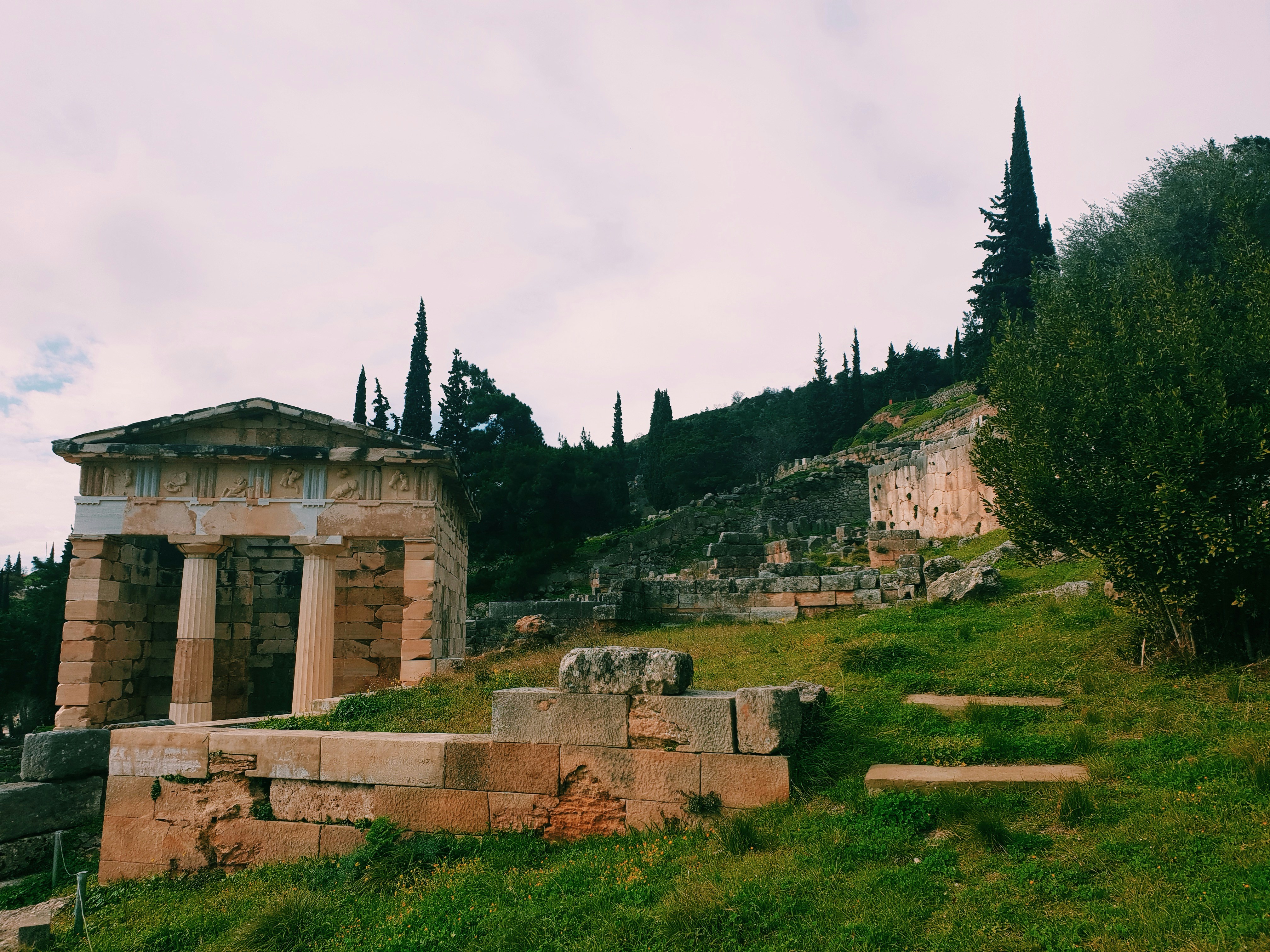 Ancient ruins featuring a small temple with columns and carved details, surrounded by greenery and trees under a cloudy sky.