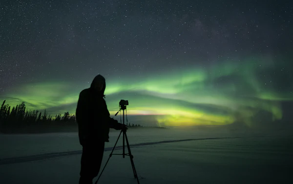 Photographer silhouetted beside a tripod on a snowy plain under a green aurora band and a starry sky