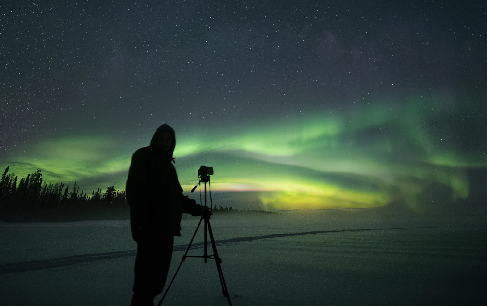 Photographer silhouetted beside a tripod on a snowy plain under a green aurora band and a starry sky