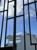 Metal window bars frame a view of a bright blue sky with scattered white clouds. In the background, parts of a white building with architectural details such as railings and a water tank are visible.