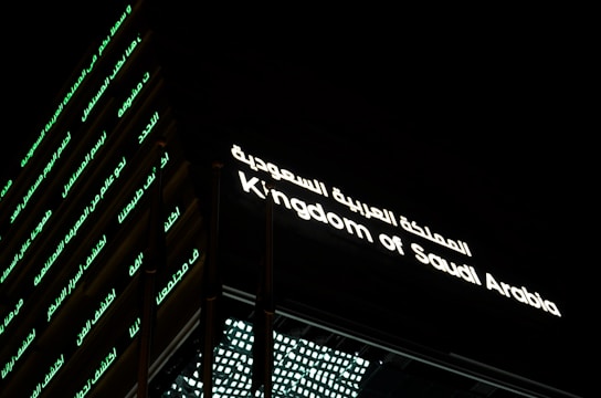 A building facade prominently displaying illuminated text. The text is in both Arabic and English, reading 'Kingdom of Saudi Arabia.' The structure's exterior features horizontal lines of green-lit Arabic script against a dark background.