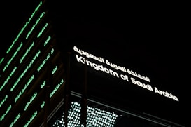 A building facade prominently displaying illuminated text. The text is in both Arabic and English, reading 'Kingdom of Saudi Arabia.' The structure's exterior features horizontal lines of green-lit Arabic script against a dark background.