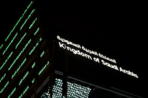 A building facade prominently displaying illuminated text. The text is in both Arabic and English, reading 'Kingdom of Saudi Arabia.' The structure's exterior features horizontal lines of green-lit Arabic script against a dark background.