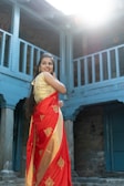 A woman wearing a vibrant red and gold saree stands in an old building with blue wooden architectural elements. She smiles softly while looking away from the camera, and sunlight streams down, adding a warm tone to the scene.