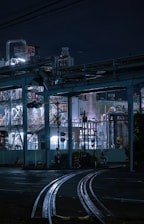 An industrial scene at night featuring a complex network of metal structures, pipes, and rail tracks. The facility is illuminated by bright artificial lights, casting shadows and highlighting the intricate details of the infrastructure. The night sky serves as a dark backdrop.