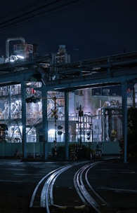 An industrial scene at night featuring a complex network of metal structures, pipes, and rail tracks. The facility is illuminated by bright artificial lights, casting shadows and highlighting the intricate details of the infrastructure. The night sky serves as a dark backdrop.