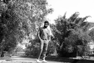 Man dressed in a sleek black suit with a relaxed yet polished look, standing in a rustic outdoor setting.