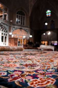 A vibrant and intricately patterned carpet is displayed prominently, with a focus on its detailed floral designs. In the background, an ornate architectural setting features arched doorways, tall windows, and warm lighting that creates a cozy atmosphere.