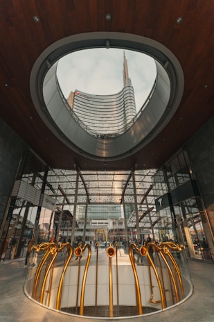 A modern architectural space featuring a view of a tall, curving skyscraper through an oval opening in the ceiling. The structure showcases contemporary design with glass walls and wooden elements. The foreground displays an art installation with several metallic pipes, golden in color, forming abstract shapes.