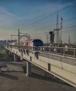 A metro train travels on an elevated track with power lines overhead. The train is modern and brightly colored, contrasting with the concrete structure. In the background, the sky is clear with various utility towers visible, indicating an urban setting.
