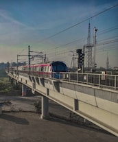 A metro train travels on an elevated track with power lines overhead. The train is modern and brightly colored, contrasting with the concrete structure. In the background, the sky is clear with various utility towers visible, indicating an urban setting.