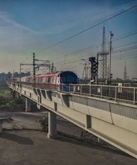 A metro train travels on an elevated track with power lines overhead. The train is modern and brightly colored, contrasting with the concrete structure. In the background, the sky is clear with various utility towers visible, indicating an urban setting.