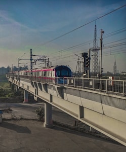 A metro train travels on an elevated track with power lines overhead. The train is modern and brightly colored, contrasting with the concrete structure. In the background, the sky is clear with various utility towers visible, indicating an urban setting.