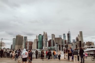 A diverse group of people stand on a boardwalk by the water, with a backdrop of a city skyline featuring numerous skyscrapers under an overcast sky.