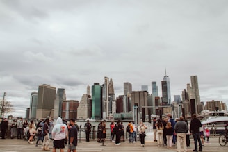 A diverse group of people stand on a boardwalk by the water, with a backdrop of a city skyline featuring numerous skyscrapers under an overcast sky.
