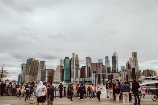 A diverse group of people stand on a boardwalk by the water, with a backdrop of a city skyline featuring numerous skyscrapers under an overcast sky.