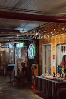 A dimly lit indoor space featuring a small shop with a colorful table covered in various small items. The wall is adorned with vibrant neon signs, one of which includes the word 'tattoo'. A person wearing a black mask and casual attire is seated nearby, glancing at something off-camera. The ceiling has an industrial look with exposed pipes, and small lights strung decoratively across.