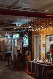 A dimly lit indoor space featuring a small shop with a colorful table covered in various small items. The wall is adorned with vibrant neon signs, one of which includes the word 'tattoo'. A person wearing a black mask and casual attire is seated nearby, glancing at something off-camera. The ceiling has an industrial look with exposed pipes, and small lights strung decoratively across.