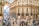 A bustling shopping arcade with a glass roof and intricate architectural details. The floor has a colorful mosaic pattern and the arcade is lined with high-end stores, including Dior and Prada. Numerous people are walking around or standing in groups.
