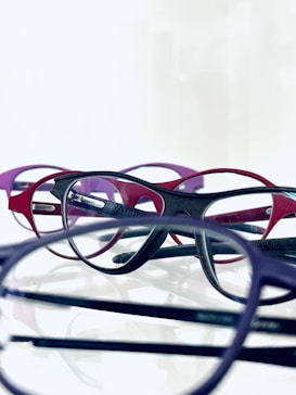 A collection of eyeglasses with frames of different colors, including red, purple, and black, are arranged closely together on a reflective surface. The glasses overlap, creating a layered effect against a white background.