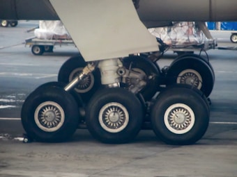 A close-up view of an airplane's landing gear with multiple wheels and mechanical components visible. The image captures a section of the aircraft on a tarmac, with other airport equipment visible in the background.