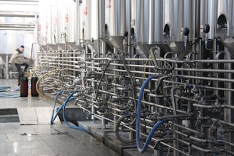 A series of large stainless steel fermentation tanks are connected by an intricate network of pipes and valves in a brewery or industrial facility. On the left side, a person in motion suggests activity and work in the space. The floor is wet, possibly from recent cleaning or maintenance.