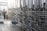 A series of large stainless steel fermentation tanks are connected by an intricate network of pipes and valves in a brewery or industrial facility. On the left side, a person in motion suggests activity and work in the space. The floor is wet, possibly from recent cleaning or maintenance.
