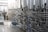 A series of large stainless steel fermentation tanks are connected by an intricate network of pipes and valves in a brewery or industrial facility. On the left side, a person in motion suggests activity and work in the space. The floor is wet, possibly from recent cleaning or maintenance.