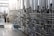 A series of large stainless steel fermentation tanks are connected by an intricate network of pipes and valves in a brewery or industrial facility. On the left side, a person in motion suggests activity and work in the space. The floor is wet, possibly from recent cleaning or maintenance.