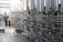 A series of large stainless steel fermentation tanks are connected by an intricate network of pipes and valves in a brewery or industrial facility. On the left side, a person in motion suggests activity and work in the space. The floor is wet, possibly from recent cleaning or maintenance.