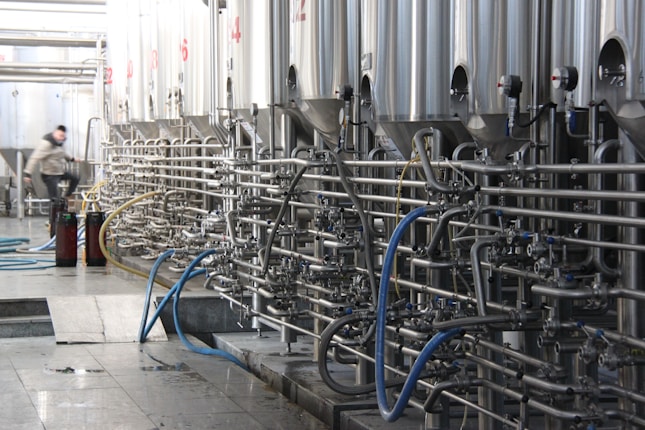 A series of large stainless steel fermentation tanks are connected by an intricate network of pipes and valves in a brewery or industrial facility. On the left side, a person in motion suggests activity and work in the space. The floor is wet, possibly from recent cleaning or maintenance.