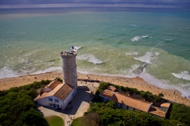 An aerial view of a coastal scene featuring a lighthouse with a French flag on top, surrounded by small buildings with red-tiled roofs. The lighthouse is situated near a sandy beach with gentle waves hitting the shore. Green vegetation borders the beach area, and a few people are visible walking around the pathways next to the buildings. The ocean water is a gradient of greenish-blue hues, and the sky above is cloudy.