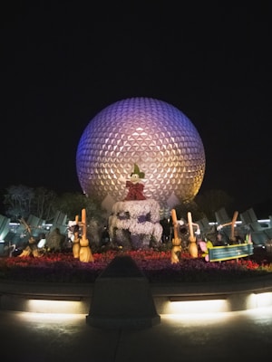 A large, illuminated geodesic sphere stands in the night, casting a gentle glow. In front of it, a vibrant garden showcases topiary sculptures resembling Disney characters, surrounded by colorful flowers. The scene is illuminated by soft lights, creating an enchanting atmosphere.