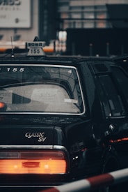 A classic black taxi cab with rain droplets visible on its surface. The taxi sign is prominently displayed on the roof, featuring Japanese characters and the number 155. The rear end of the car includes a large red taillight and the words 'CLASSIC SV' are emblazoned near the trunk.