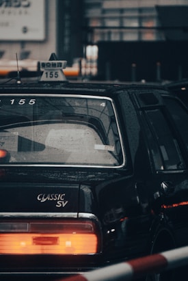 A classic black taxi cab with rain droplets visible on its surface. The taxi sign is prominently displayed on the roof, featuring Japanese characters and the number 155. The rear end of the car includes a large red taillight and the words 'CLASSIC SV' are emblazoned near the trunk.