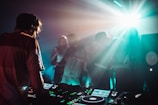 A DJ is working on a music console while a crowd of people dances in a dimly lit room with vibrant light effects. The atmosphere is energetic, with beams of light illuminating the foggy air and creating a dynamic, club-like setting.