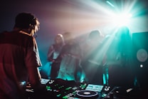 A DJ is working on a music console while a crowd of people dances in a dimly lit room with vibrant light effects. The atmosphere is energetic, with beams of light illuminating the foggy air and creating a dynamic, club-like setting.