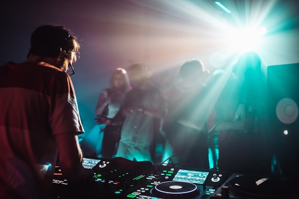 A DJ is working on a music console while a crowd of people dances in a dimly lit room with vibrant light effects. The atmosphere is energetic, with beams of light illuminating the foggy air and creating a dynamic, club-like setting.