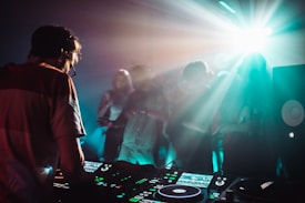 A DJ is working on a music console while a crowd of people dances in a dimly lit room with vibrant light effects. The atmosphere is energetic, with beams of light illuminating the foggy air and creating a dynamic, club-like setting.