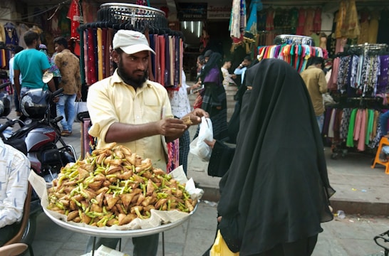 A street vendor stands behind a stall filled with crispy food items, possibly samosas, topped with green chilies. He is handing them to a customer dressed in a dark, full-length veil. The backdrop includes colorful fabrics and garments displayed on racks, with several people browsing and walking around.