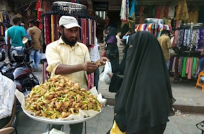 A street vendor stands behind a stall filled with crispy food items, possibly samosas, topped with green chilies. He is handing them to a customer dressed in a dark, full-length veil. The backdrop includes colorful fabrics and garments displayed on racks, with several people browsing and walking around.