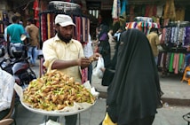 A street vendor stands behind a stall filled with crispy food items, possibly samosas, topped with green chilies. He is handing them to a customer dressed in a dark, full-length veil. The backdrop includes colorful fabrics and garments displayed on racks, with several people browsing and walking around.