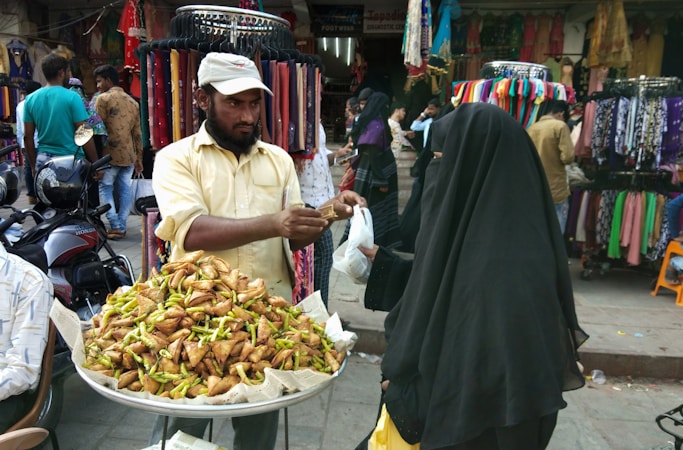 A street vendor stands behind a stall filled with crispy food items, possibly samosas, topped with green chilies. He is handing them to a customer dressed in a dark, full-length veil. The backdrop includes colorful fabrics and garments displayed on racks, with several people browsing and walking around.