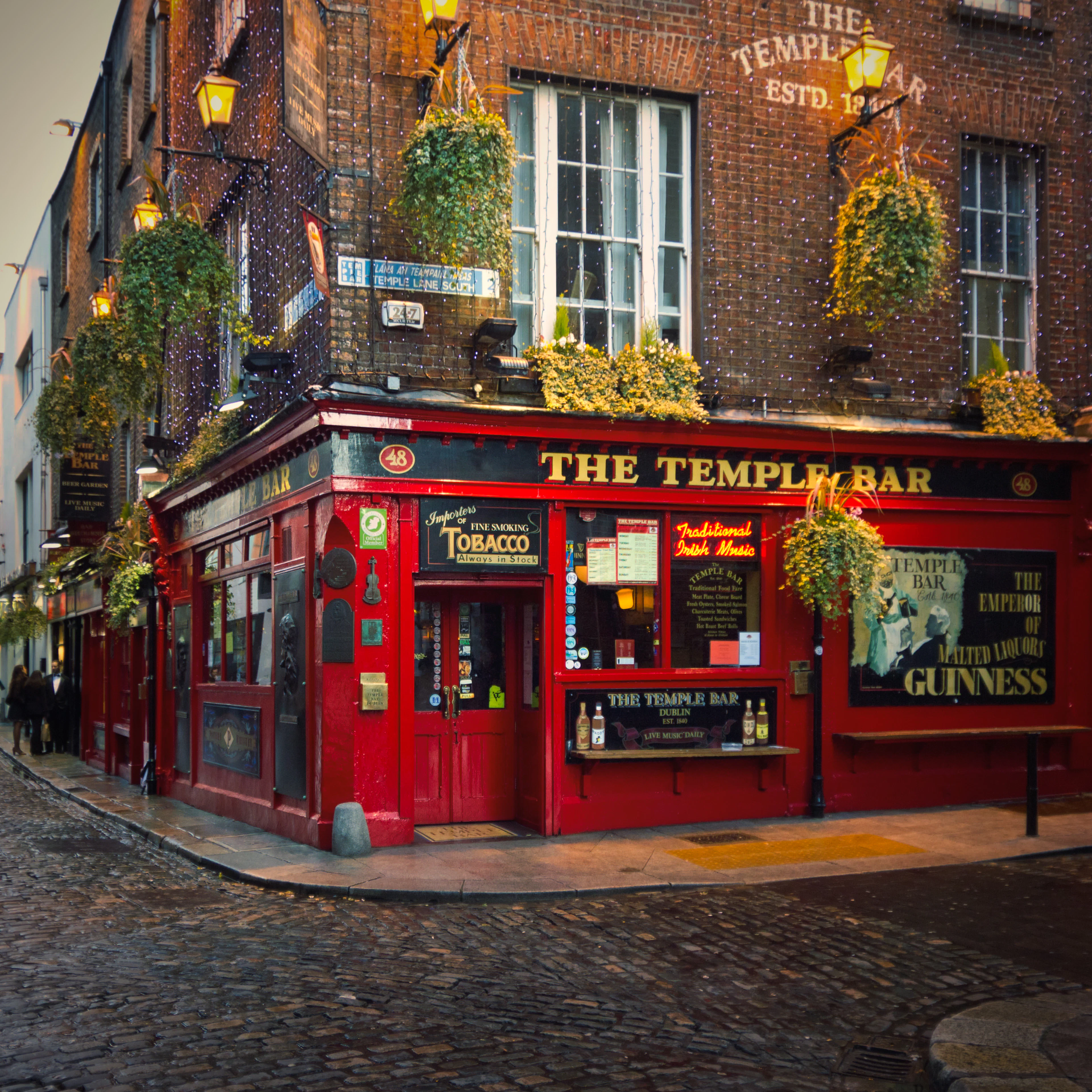The red facade of The Temple Bar pub on a wet cobblestone corner, hung with flower baskets and string lights.