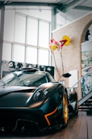 A sleek, high-performance sports car with a glossy black finish and distinct yellow accents is parked indoors. The car is positioned at an angle that showcases its aerodynamic design, headlights, and vibrant wheels. The background includes glass windows, architectural elements, and a partially visible staircase. A striking red and yellow artistic installation hangs from the ceiling.