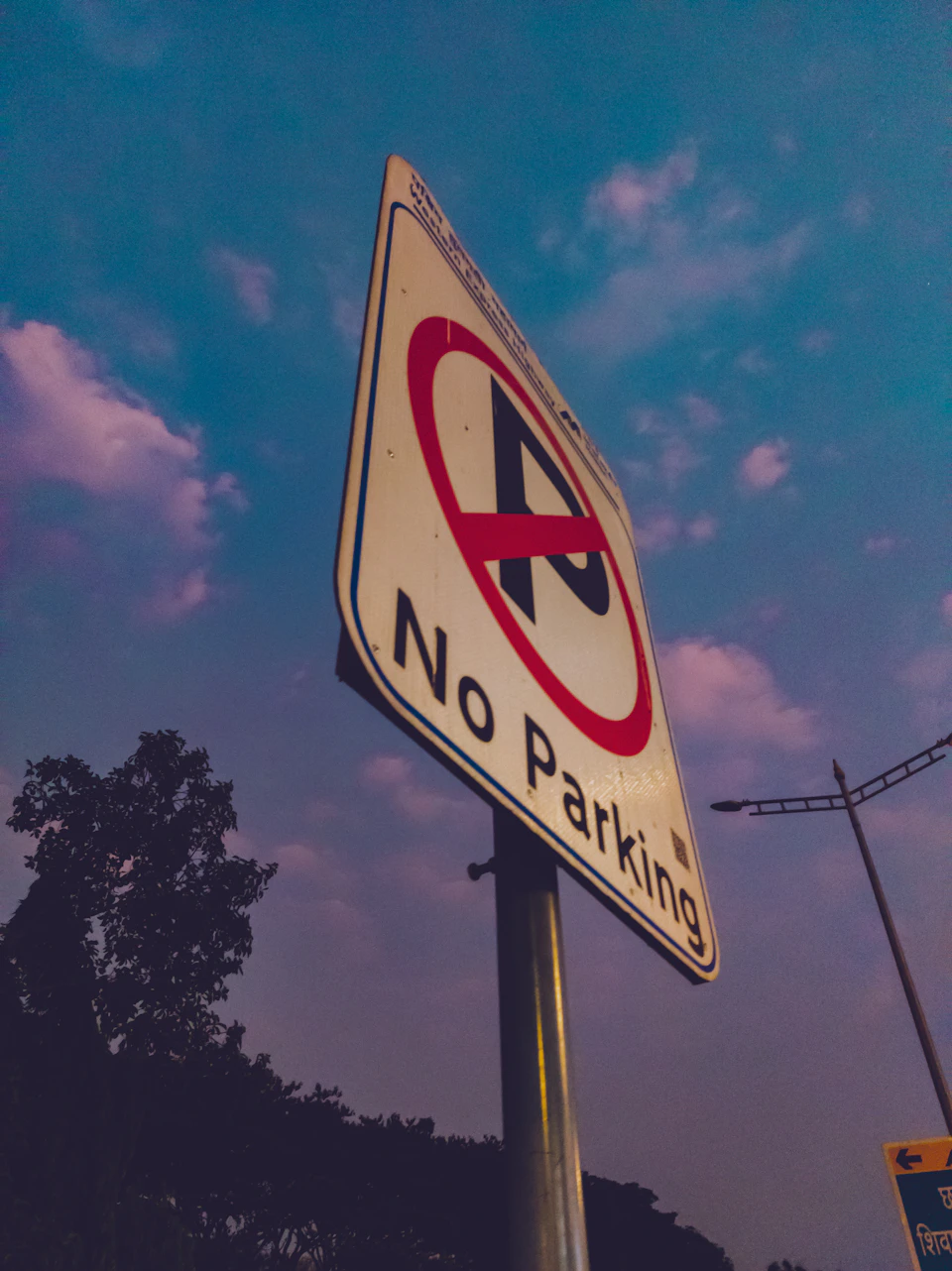 The image shows a street sign mounted on a metal pole, displaying a "No Parking" message. The sign is white with black text and features an exclamation mark at the top. Below the sign, there are two street lights visible in the background.